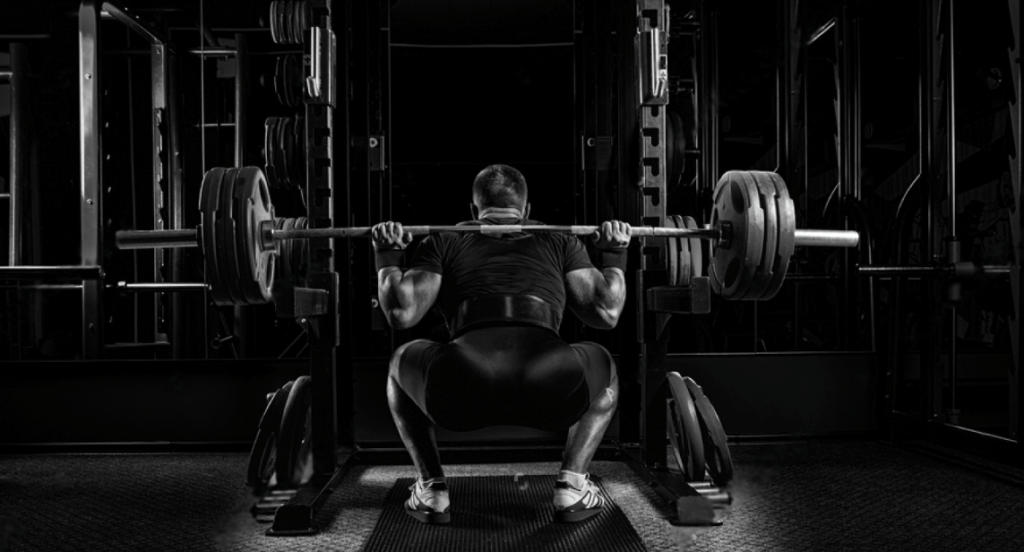 Professional athlete sits with a barbell on his shoulders and prepares to stand with her. View from the back